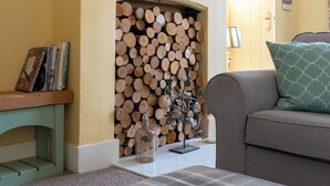 A living room with a fireplace, a round mirror, a stack of firewood, a sofa, a coffee table, and a bookshelf.