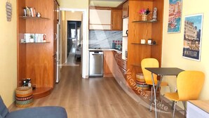 A compact living space with a kitchenette, wooden cabinets, a dining area with yellow chairs, and a sofa. - T2 SPACIEUX, LOGGIA ET ACCES WIFI (Sète)
