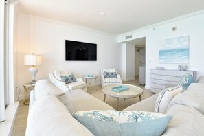 Sala de estar com um sofá branco, duas poltronas, uma mesa de centro e uma mesa lateral com abajur. Há um quadro na parede e vista para o oceano pela janela.
