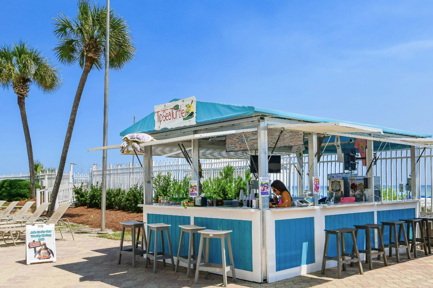 Un quiosco de comida en la playa con un letrero que dice 'TipSeaTurtle', con mostrador, taburetes y un panel de menú.