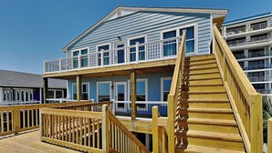 Ein mehrstöckiges Gebäude direkt am Strand mit einer Holzterrasse und einer Treppe, die zum oberen Stockwerk führt.