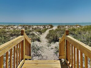 Ein hölzerner Steg führt zu einem Strand mit Dünen und spärlicher Vegetation.