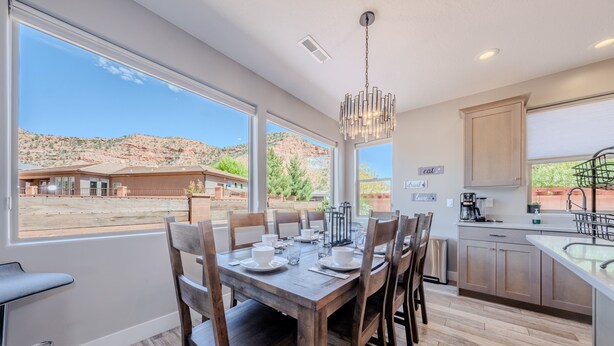 A dining area with a wooden table set for six, large windows offering a view of a desert landscape, and a modern kitchen with wooden cabinets and a stainless steel sink.