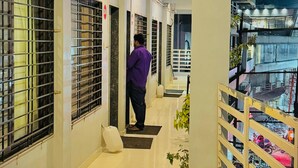 A corridor with tiled floors, a person in a purple shirt, and a railing with a view of parked motorcycles. - Hotel Sai Navsari (Jalalpore)