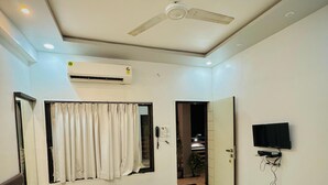 A modern hotel room with a bed, ceiling fan, air conditioning unit, television, and a view into the adjacent room. - Hotel Sai Navsari (Jalalpore)