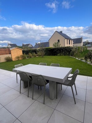 A patio with a rectangular table and six chairs, surrounded by a well-maintained lawn and a house in the background. - Villa Bambou, 5 minutes from Saint-Malo—a new and quiet home! (Saint-Jouan-des-Guérets)