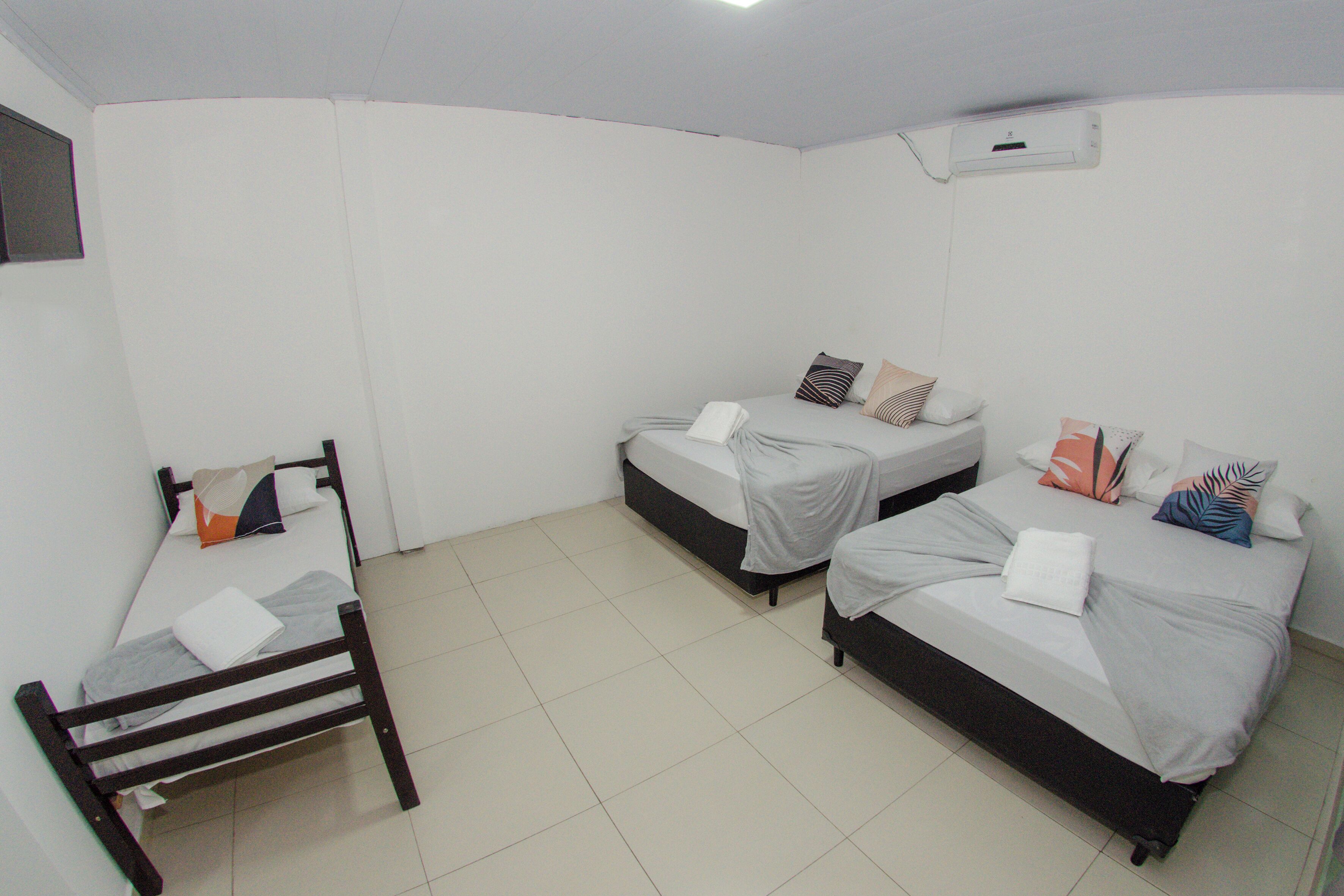 Una habitación con tres camas, cada una con diseños de almohadas distintos, una silla de madera y un equipo de aire acondicionado blanco en la pared.