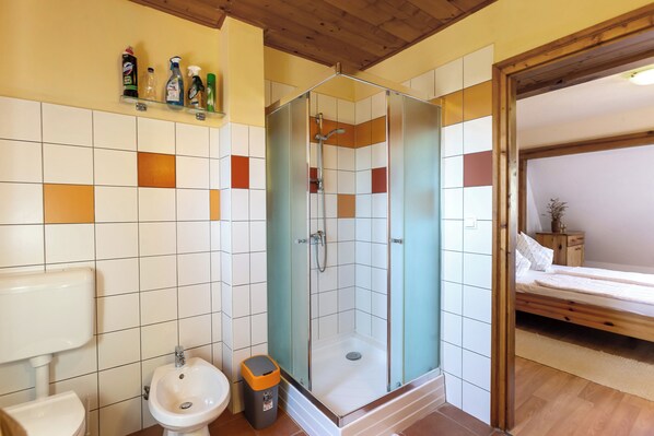 Ein Badezimmer mit Dusche, Waschbecken und Toilette. Im angrenzenden Raum befindet sich ein Bett.