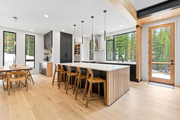 A modern kitchen with a central island, wooden chairs, and large windows offering a view of the forest.