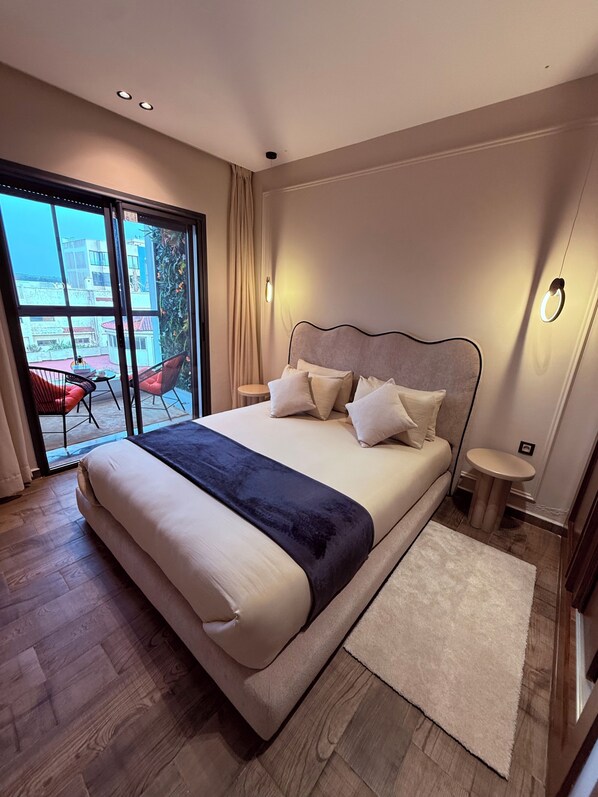 A modern bedroom with a large bed, a small bedside table, a nightstand, a sliding glass door to a balcony, and a view of buildings outside.