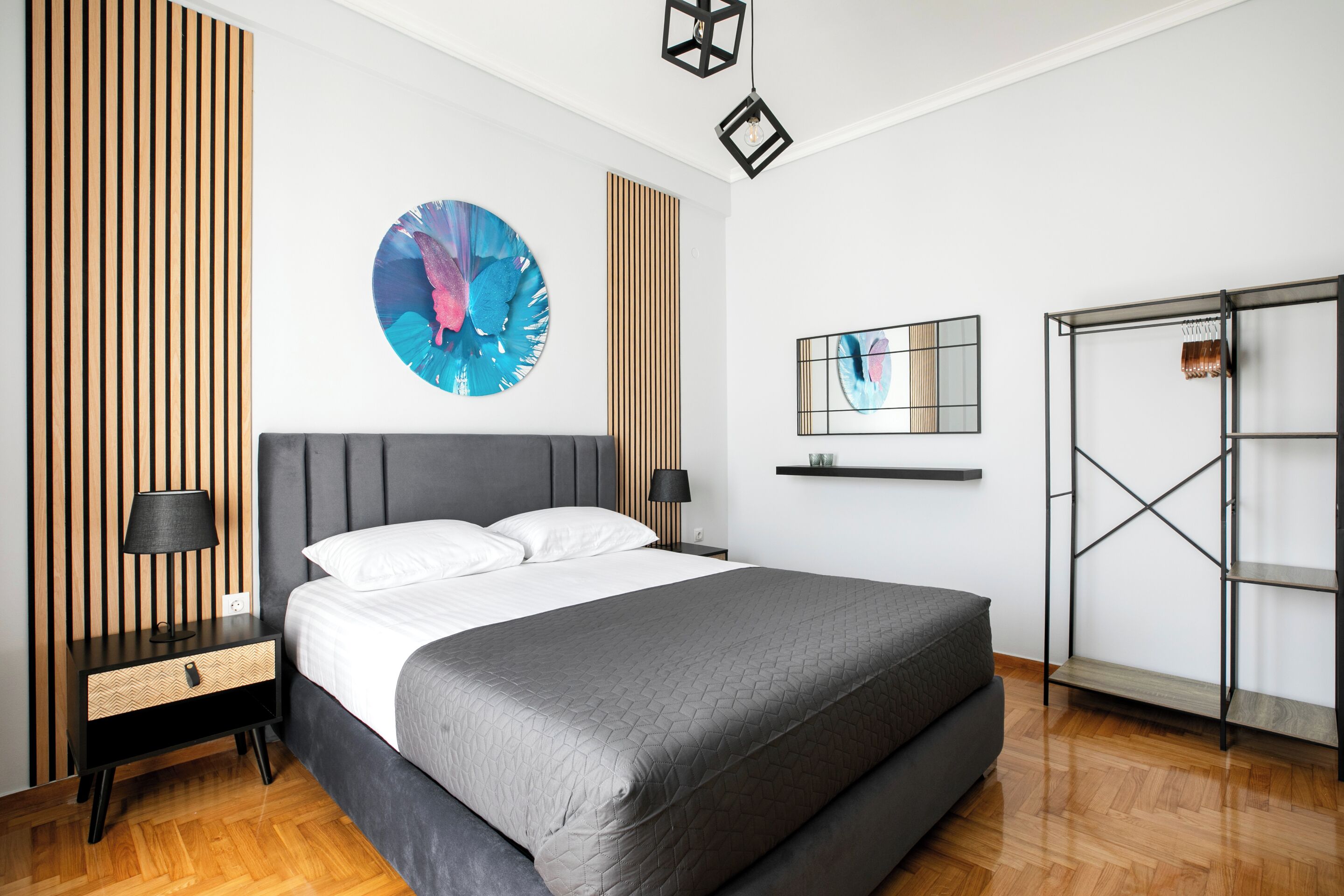 A modern bedroom with a large bed, a nightstand with a lamp, a wall-mounted shelf, and a metal shelving unit.