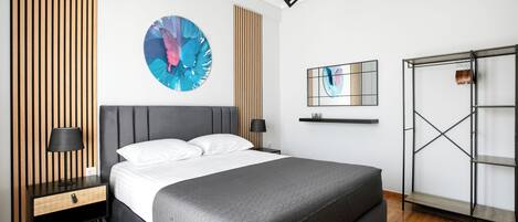 A modern bedroom with a large bed, a nightstand with a lamp, a wall-mounted shelf, and a metal shelving unit.