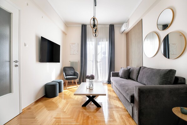 Een moderne woonkamer met een grijze bank, een salontafel, een aan de muur bevestigde televisie en een houten vloer.