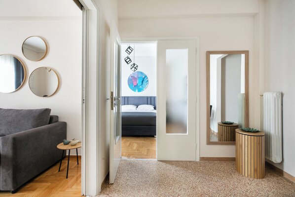 A modern hotel room with a grey sofa, a small round table with decorative items, a bed with a grey headboard, a large mirror, and a wooden side table.