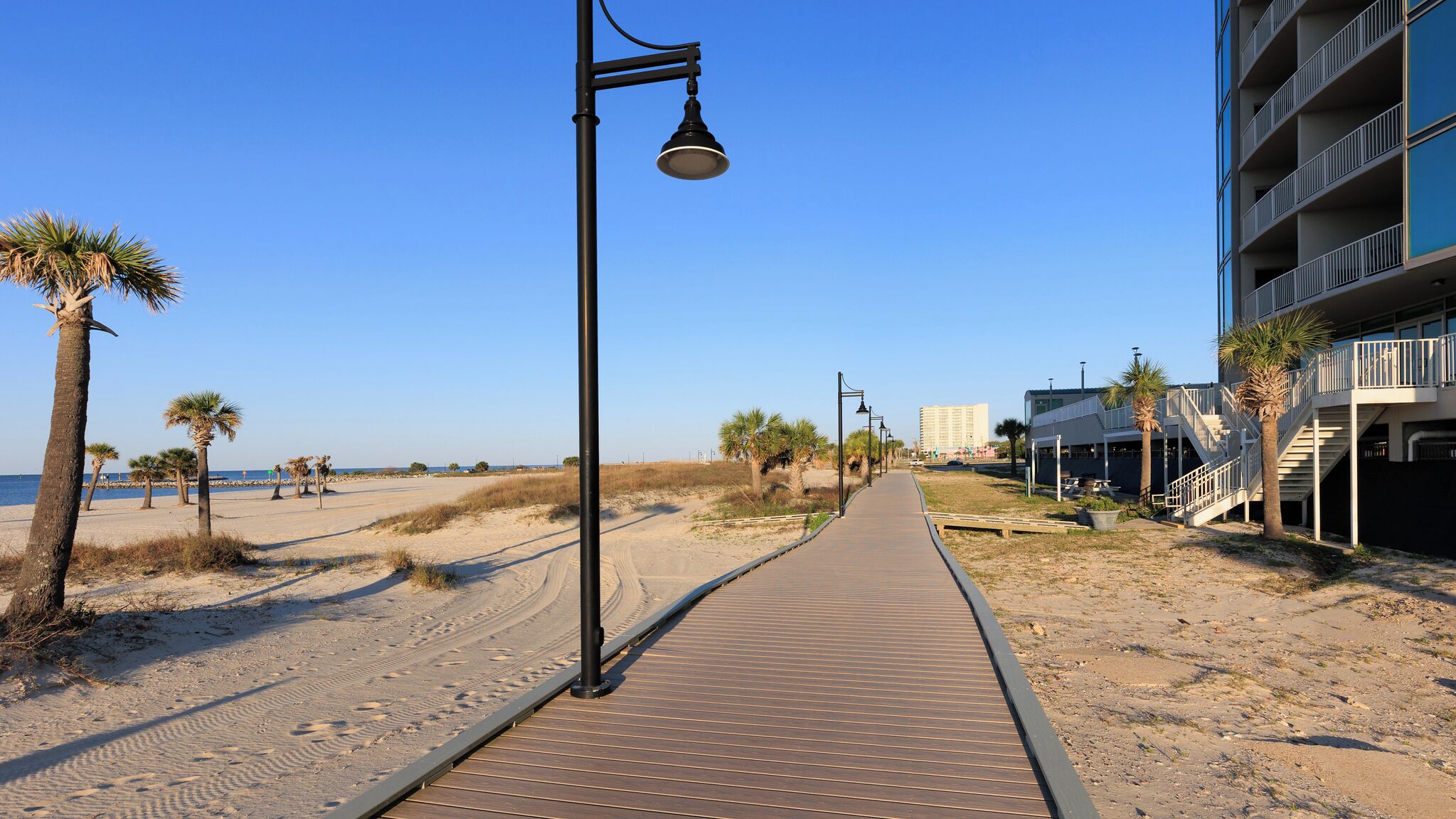 A wooden boardwalk along a sandy beach with palm trees and a building with balconies.