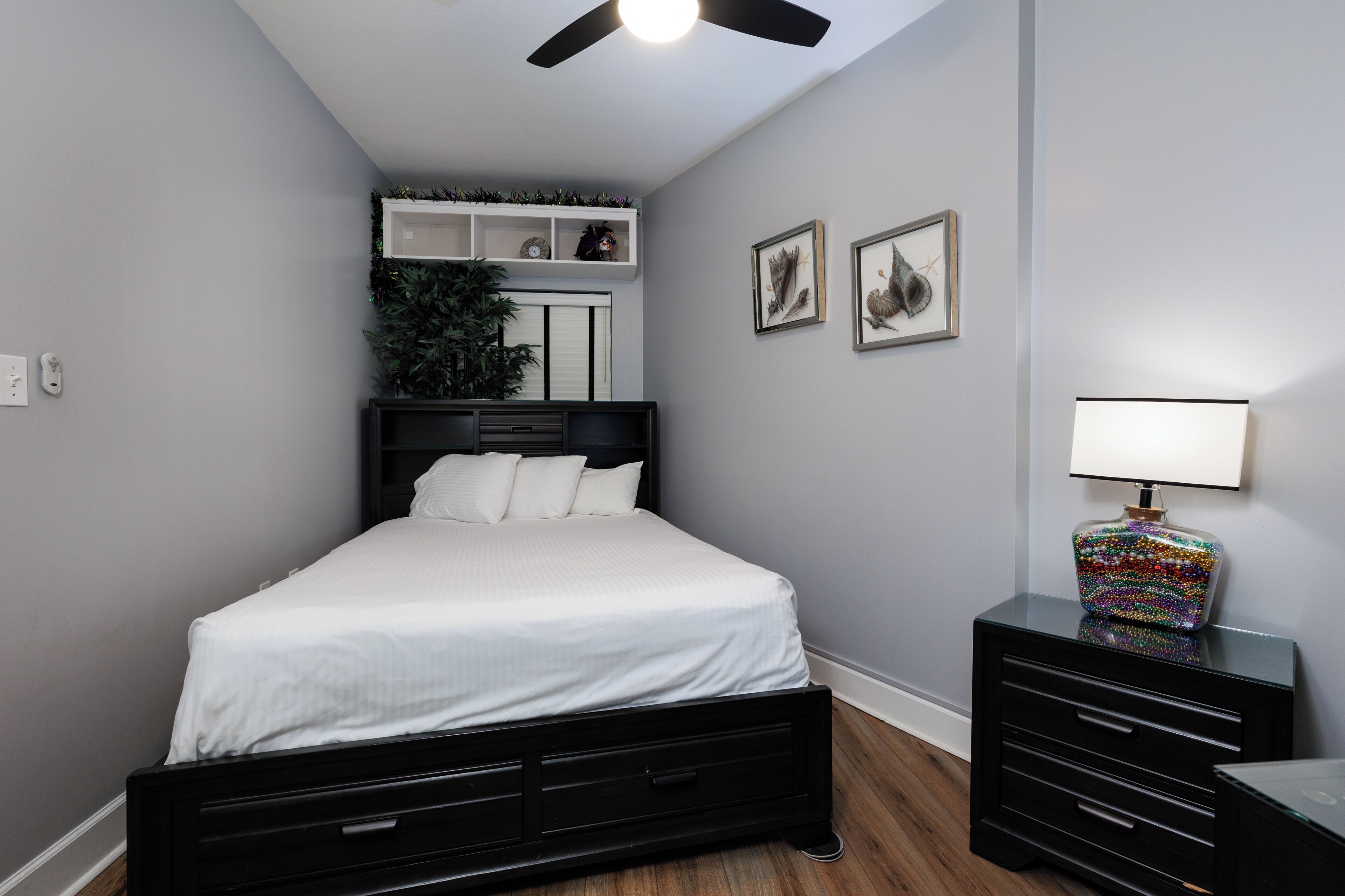 Une chambre à coucher comprenant un lit, une commode, une table de chevet, un ventilateur de plafond, une étagère fixée au mur avec des objets décoratifs, et des tableaux encadrés accrochés au mur.