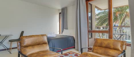 Hotelkamer met een bed, twee leren fauteuils, een houten tafel en een balkon met uitzicht op palmbomen.