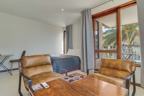 Ein Hotelzimmer mit einem Bett, zwei Ledersesseln, einem Holztisch und einem Balkon mit Blick auf Palmen.