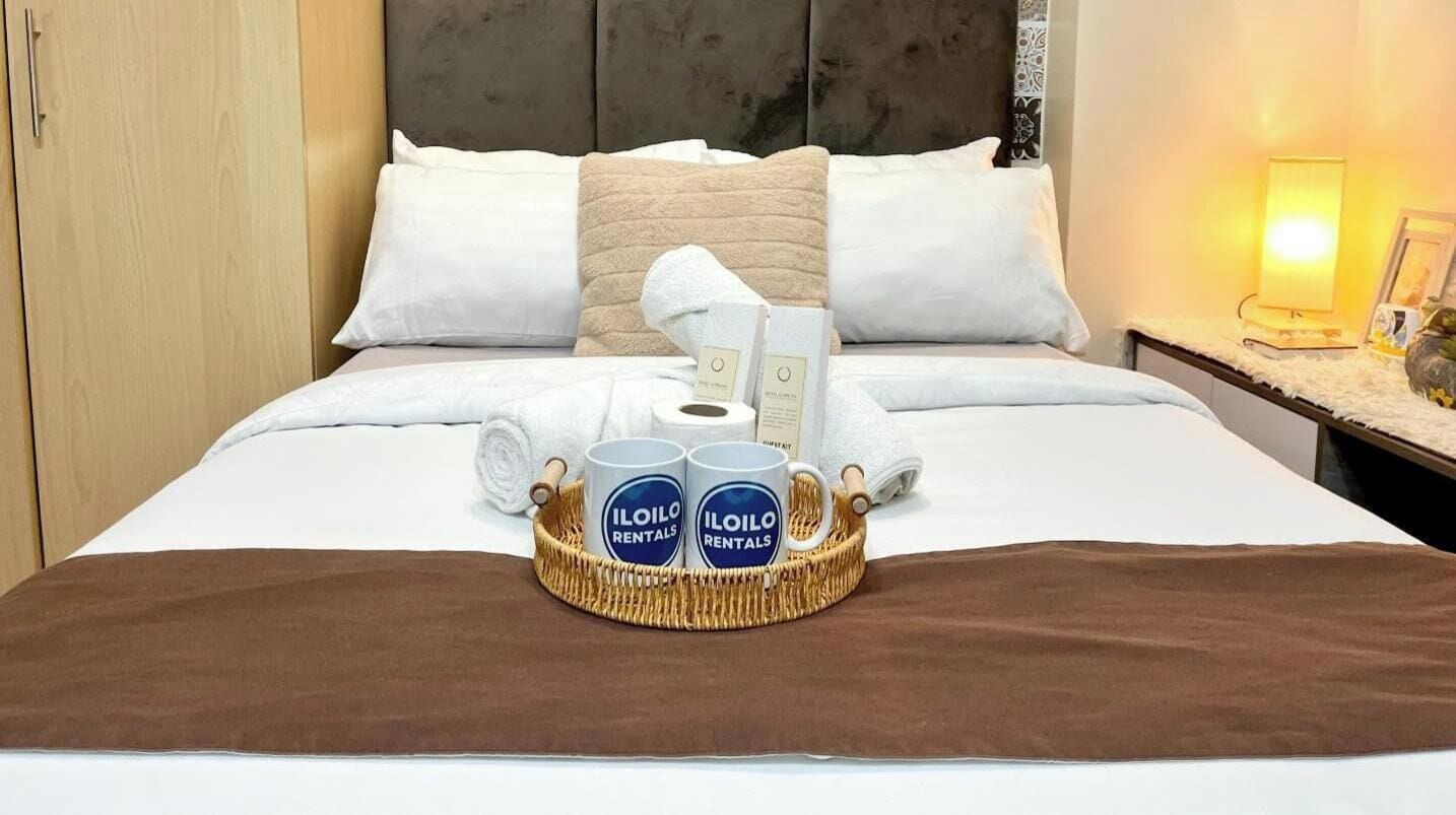 A neatly made bed with a brown bedspread, two white pillows, and a headboard. A wicker tray with two mugs labeled 'ILOILO RENTALS' and a rolled towel is placed on the bed.