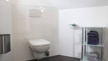 Une salle de bain moderne équipée de toilettes, d’une poubelle et d’une étagère en verre où sont pliées des serviettes.
