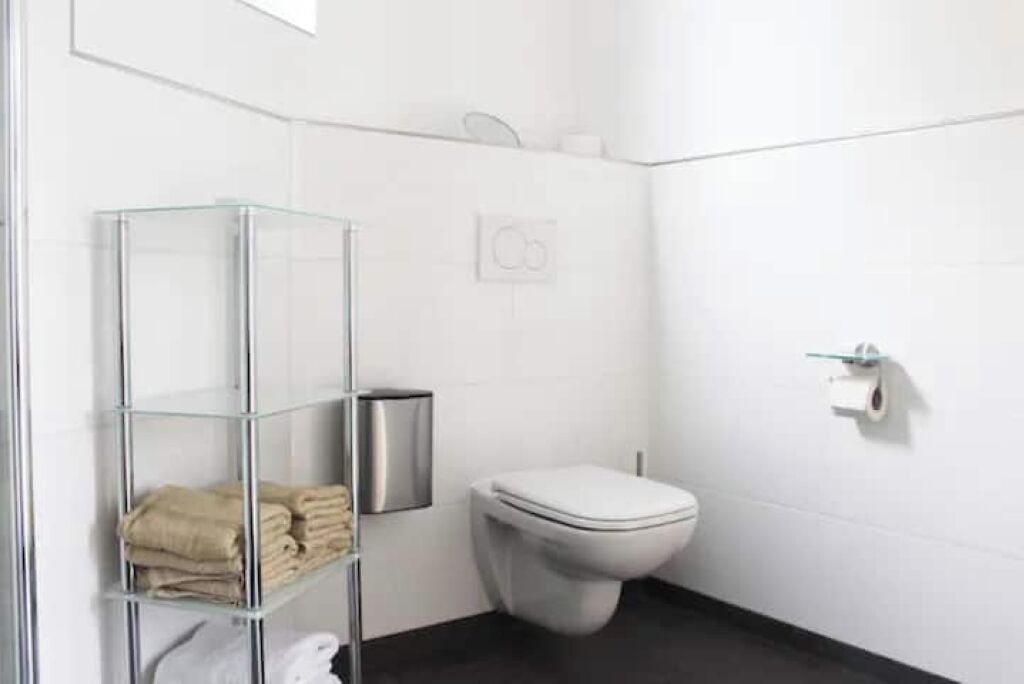 A modern bathroom with a glass shelf holding towels, a toilet, and a trash bin.