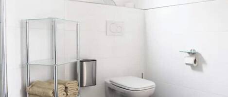 A modern bathroom with a glass shelf holding towels, a toilet, and a trash bin.