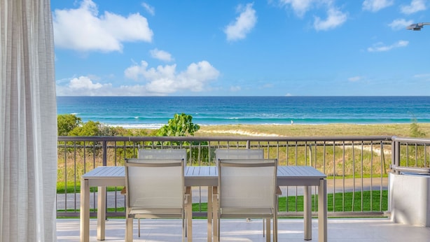 A balcony with three chairs and a table, overlooking a beach with the ocean and a clear sky.