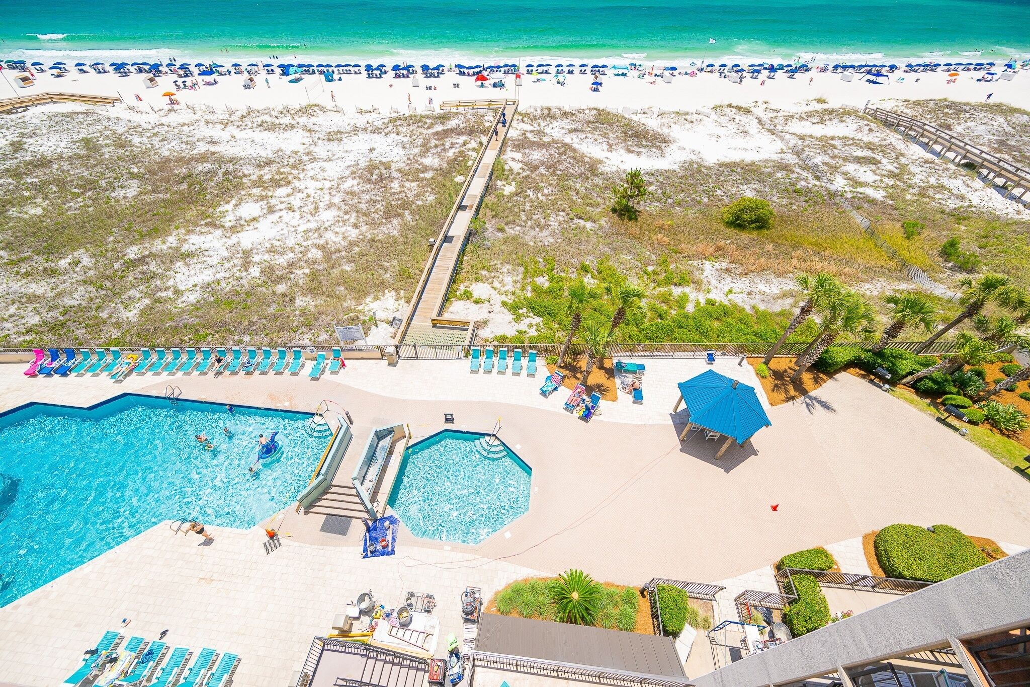 A beach resort with a swimming pool, beach umbrellas, and a wooden walkway leading to the beach.