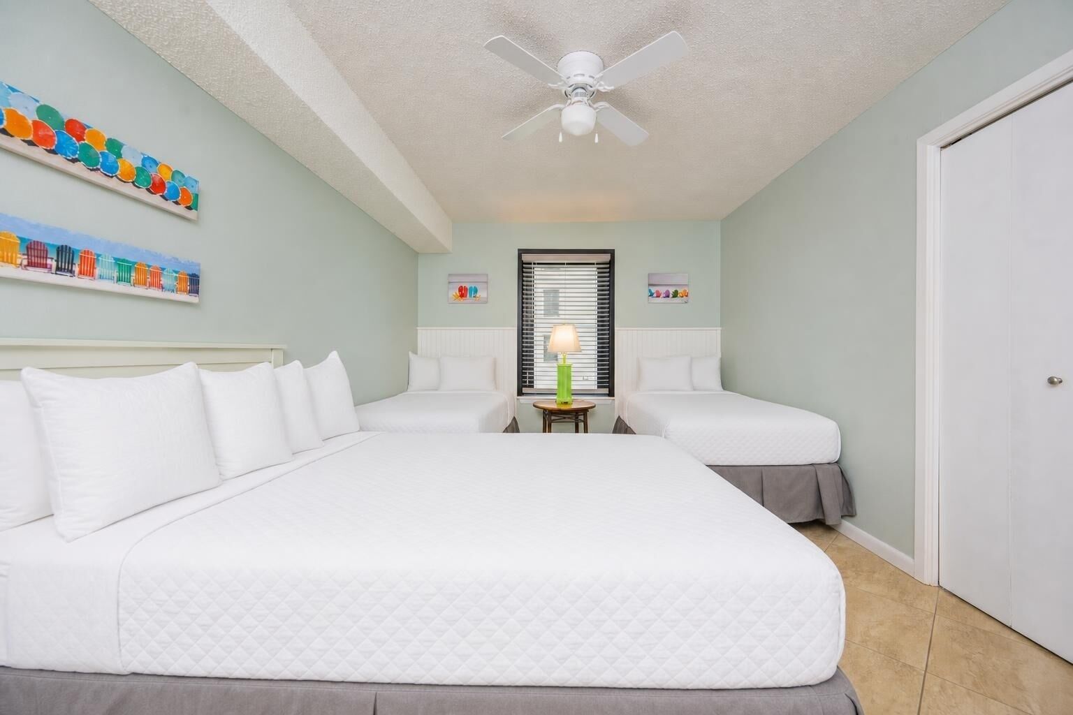 A hotel room with a large bed, two smaller beds, a ceiling fan, a window with blinds, and artwork on the walls.
