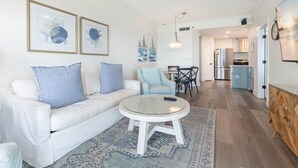 Ein modernes Wohnzimmer mit einem weißen Sofa, blauen Kissen, einem Glastisch und einem gemusterten Teppich. Es gibt eine Essecke mit Tisch und Stühlen, und im Hintergrund eine Küche.