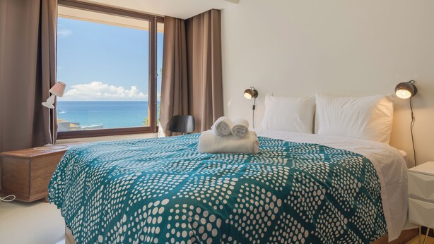 A hotel room with a large bed, a view of the sea, and a nightstand with a lamp. - Carlos Apartment (Funchal)