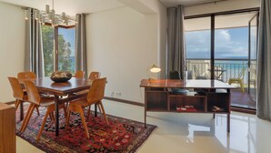 A dining area with a wooden table, chairs, and a rug. There is a chandelier above the table and a desk with a lamp. Large windows with curtains are visible, offering a view of the outdoors. - Carlos Apartment (Funchal)
