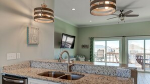 Una cocina con encimeras de granito, pileta de acero inoxidable, muebles blancos, ventilador de techo y un amplio ventanal con vista a una terraza.