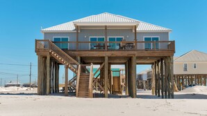 Una casa de madera de dos pisos con un balcón en el nivel superior, construida sobre pilotes sobre una playa arenosa.