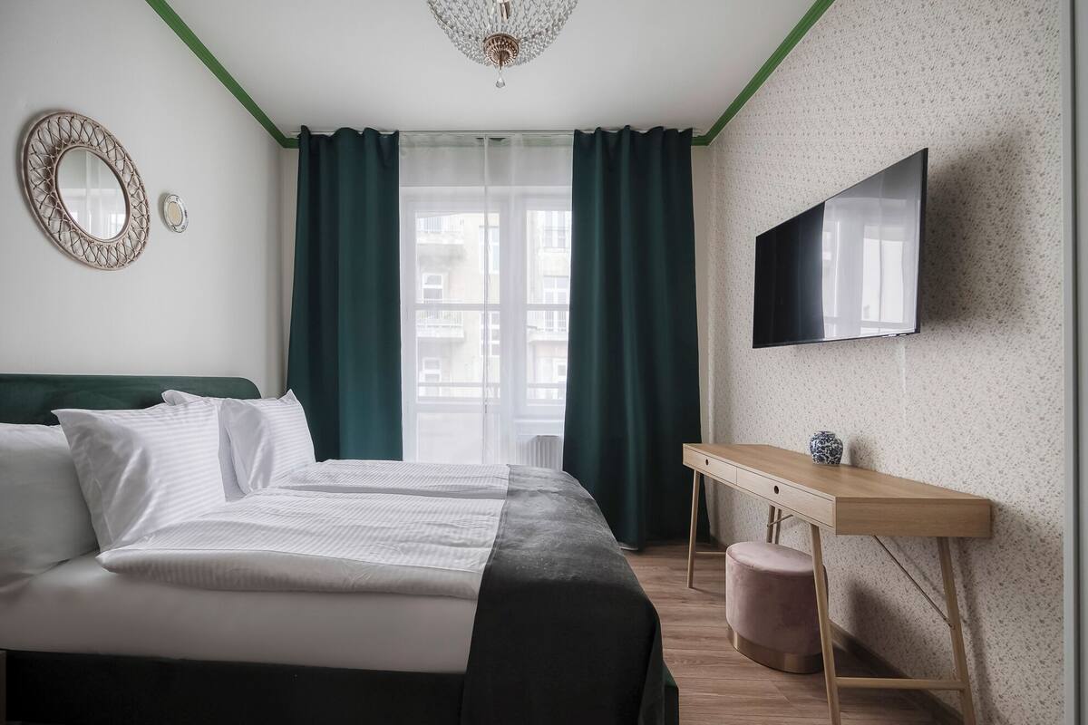 A modern hotel room with a bed, a desk, a television, and a view of the city through large windows.