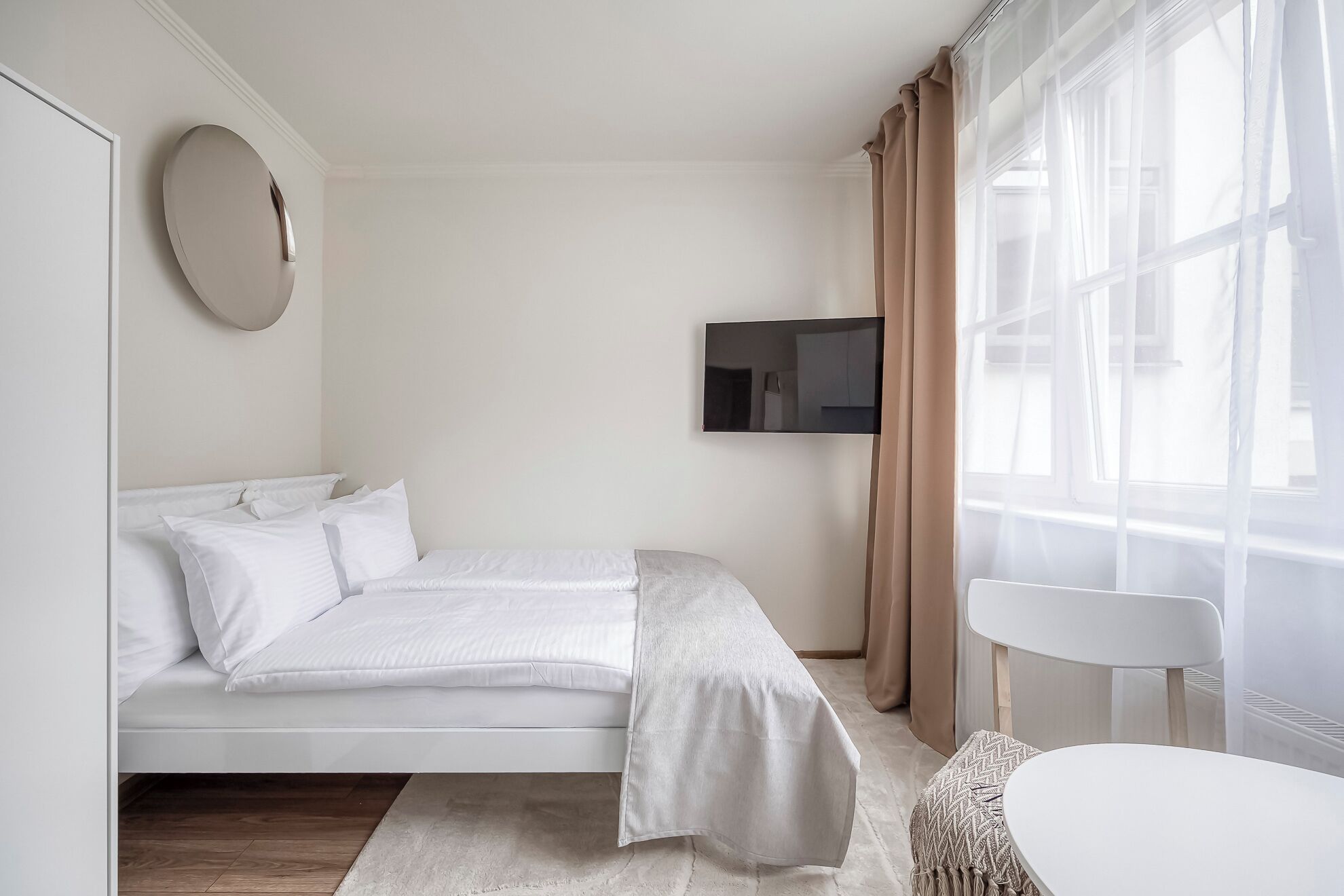Camera d'albergo minimalista con un letto, una TV a muro, uno specchio rotondo e una finestra con tende leggere.