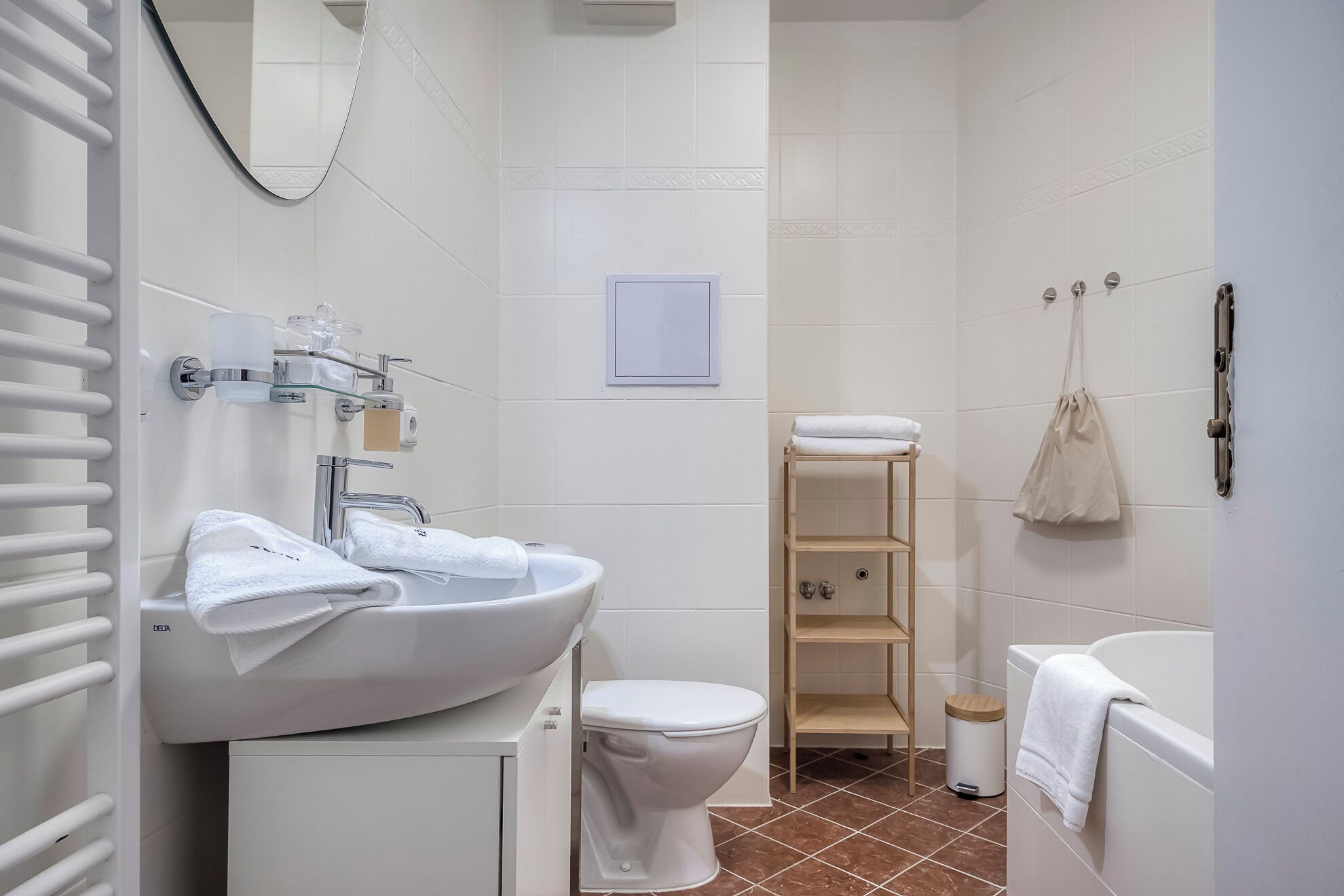 Un baño moderno con lavamanos, inodoro y bañera blancos. Se observa un perchero con toallas, un dispensador de jabón de vidrio y una pequeña estantería.