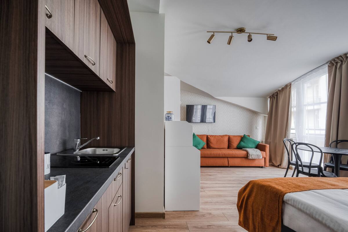 A modern kitchen with dark wood cabinets, a black countertop, and a stainless steel sink. There is a white sofa with green and orange cushions in the living area, and a bed with an orange blanket.