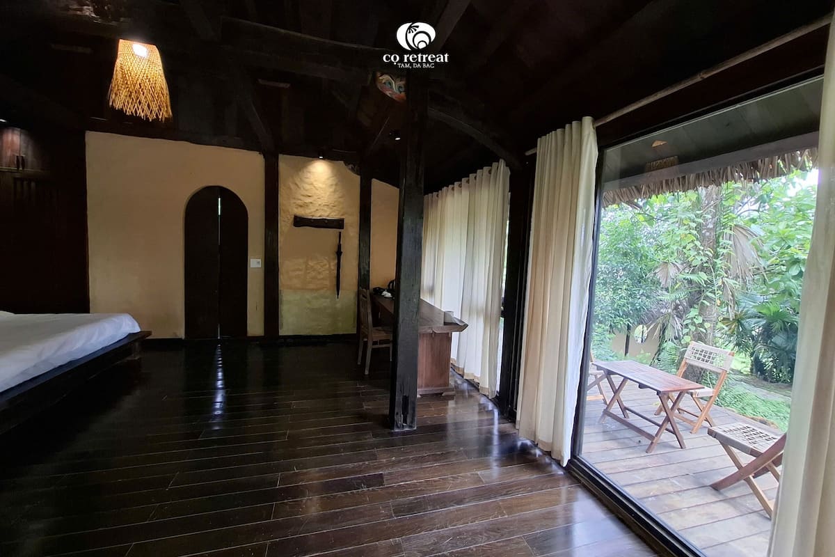 A room with a wooden floor, a bed, a desk, and a view of greenery through a glass door.