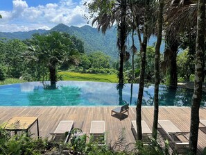 A infinity pool with wooden deck and lounge chairs, surrounded by lush greenery and mountains. - Co Retreat Da Bac (Quy Duc)