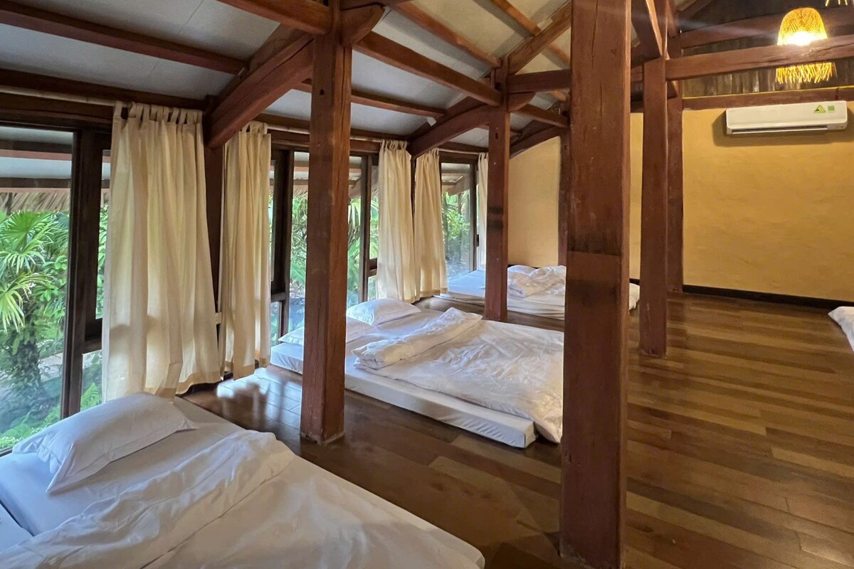 A room with wooden beams, two beds, a ceiling fan, and a view of greenery outside.