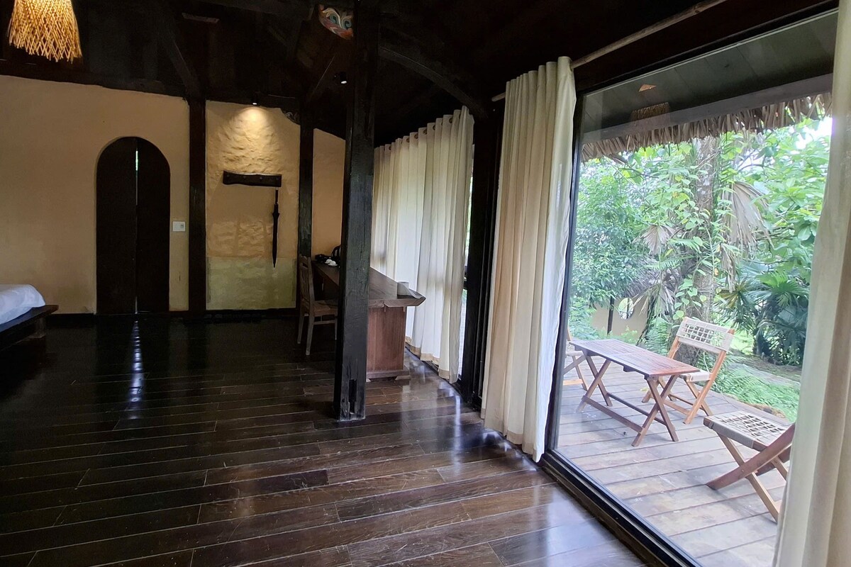 A room with a wooden floor, a bed, a desk, and a view of greenery through a glass door.