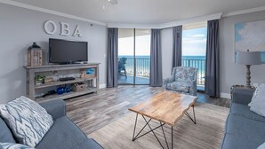 Una sala de estar con televisor, una mesa de centro de madera, sofás grises y un balcón con vista al mar.