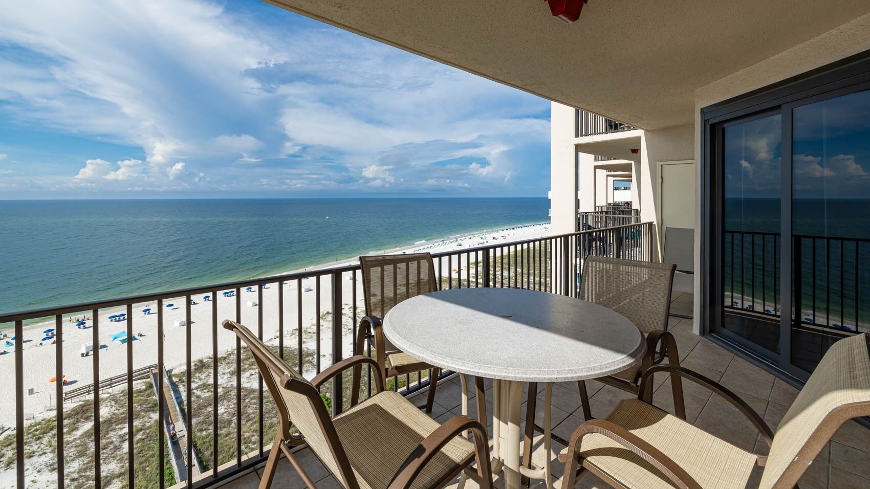 A balcony with a round table and chairs, overlooking a beach with umbrellas and a clear view of the ocean.