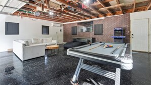 Une pièce avec une table de billard, un canapé, un fauteuil poire, une télévision et un mur en briques.