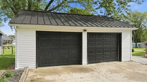 Uma garagem térrea com duas portas de garagem pretas, telhado de metal e calçada de concreto.