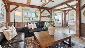 Un intérieur rustique avec des poutres apparentes, un canapé en cuir, une table basse en bois et une grande fenêtre équipée de stores.