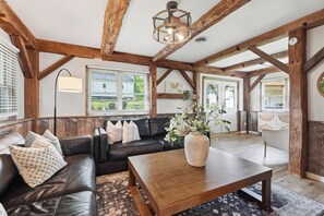 Un intérieur rustique avec des poutres apparentes, un canapé en cuir, une table basse en bois et une grande fenêtre équipée de stores.