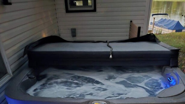 Un bain à remous doté d’un siège intégré et de marches, entouré d’une maison avec une fenêtre et d’une zone couverte en arrière-plan.