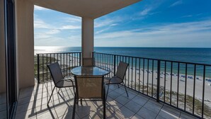 A balcony with a glass table and chairs, overlooking a beach with umbrellas and a clear sky. - Phoenix East 1203 2 Bedroom Condo (Orange Beach)